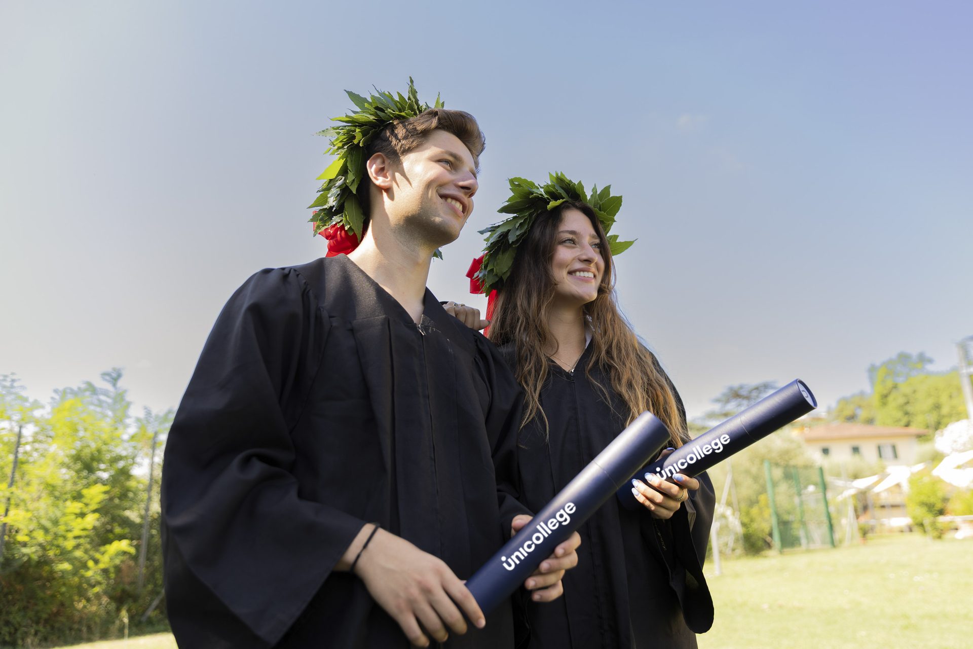 Two Unicollege graduates with laurel wreaths and diploma tubes on the campus lawn